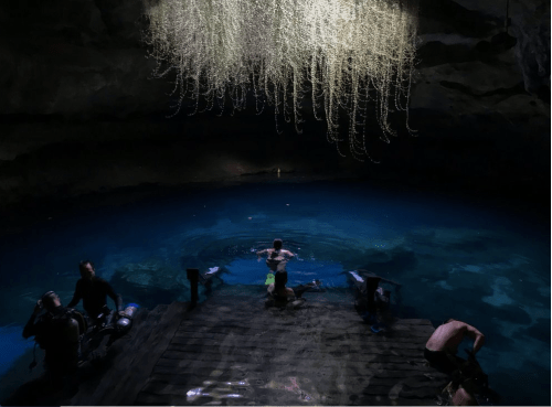 A dimly lit cave with a clear blue pool, featuring divers preparing and a striking hanging formation above.