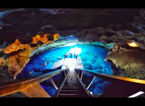 A wooden staircase leads down to a bright blue underground cave with people exploring the water.