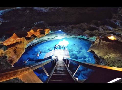 A wooden staircase leads down to a bright blue underground pool surrounded by rocky cave walls.