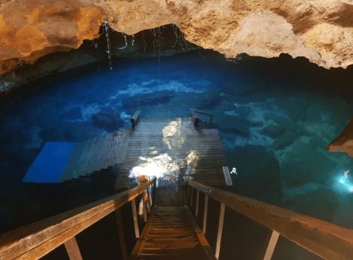 A wooden staircase leads down to a serene, blue underground pool surrounded by rocky cave walls.