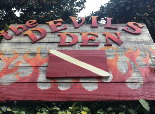 Sign for "The Devil's Den" with red letters and a white dive flag, surrounded by flames and greenery.