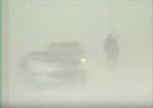 A person stands in a snowstorm near a car, surrounded by heavy snow and low visibility.