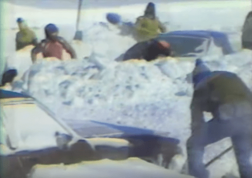 People digging out cars from deep snow during a winter storm. Snow piles surround the vehicles.