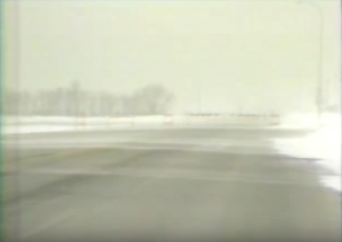 A snowy, desolate road with low visibility and trees in the background, creating a wintry scene.