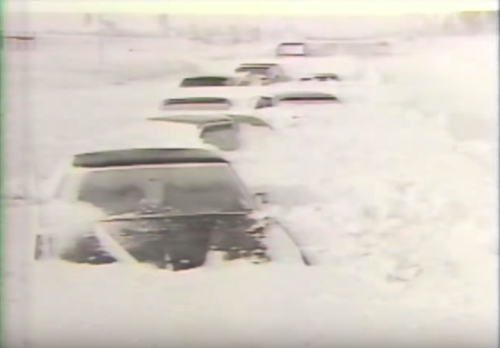 A line of cars partially buried in snow, with only the tops visible, on a snowy road.