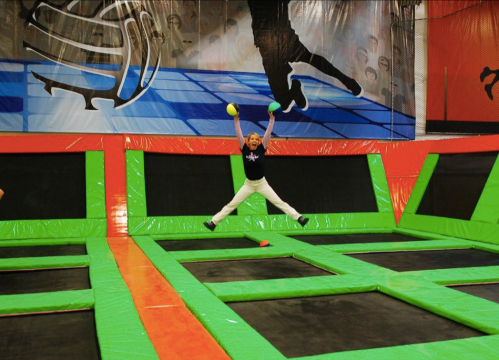 A child jumps joyfully on a trampoline, holding two balls, in a colorful indoor trampoline park.