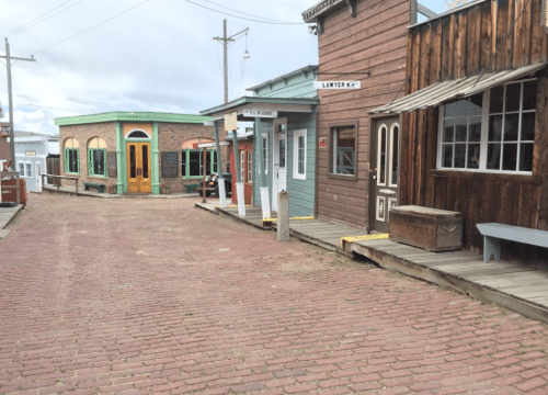 A rustic street lined with colorful wooden buildings and brick pavement, evoking a historic town atmosphere.