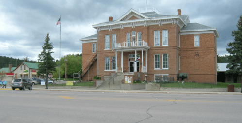 Historic brick building with a balcony, surrounded by trees and a street, under a cloudy sky. American flag visible.