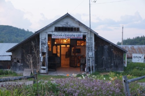A rustic barn with a sign reading "Museum of Everyday Life," surrounded by greenery and flowers.