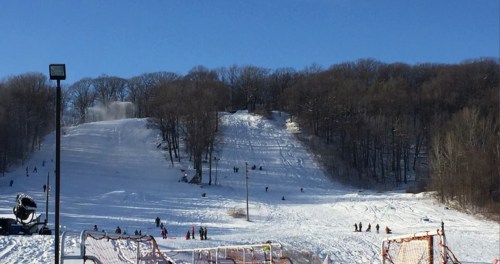 A snowy hill with people sledding and playing in the snow, surrounded by bare trees under a clear blue sky.