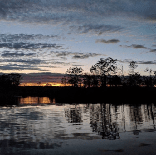 A serene sunset over a calm river, with trees silhouetted against a colorful sky and reflections on the water.