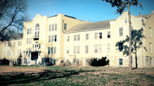 A large, historic yellow building with multiple windows and a front entrance, surrounded by grass and trees.