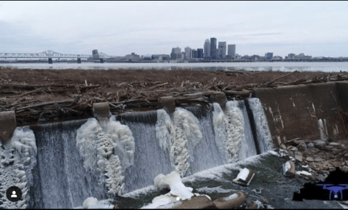 A frozen waterfall cascades over a dam, with a city skyline in the background and debris along the riverbank.