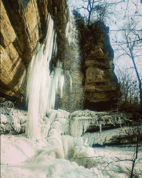 A frozen waterfall cascading down a rocky cliff, surrounded by icy formations and bare trees.