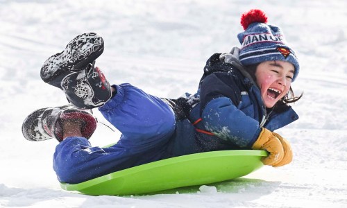 A joyful child sledding down a snowy hill, wearing a blue jacket, colorful hat, and gloves, with a big smile.