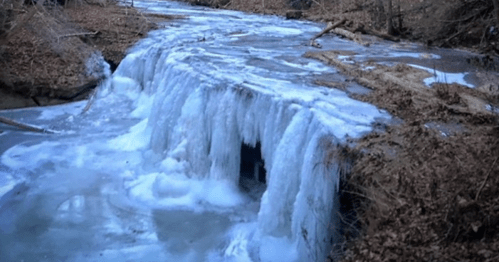 A frozen waterfall cascading over rocks, surrounded by bare trees and a winter landscape.