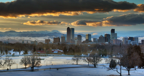 A winter sunset over a city skyline, with snow-covered ground and mountains in the background.