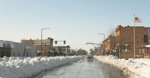A snowy street scene with a clear road, lined by buildings and an American flag, under a bright blue sky.