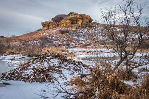 A rocky outcrop rises above a snowy landscape with a frozen pond and sparse vegetation in winter.