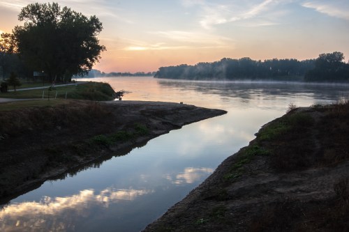 A serene river at sunrise, reflecting soft colors in the water, surrounded by trees and misty banks.
