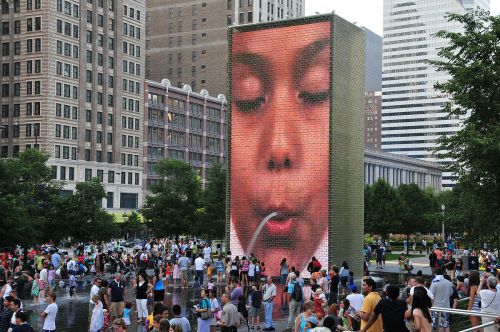 A large digital fountain displays a close-up of a child's face, with people playing in the water below in an urban park.