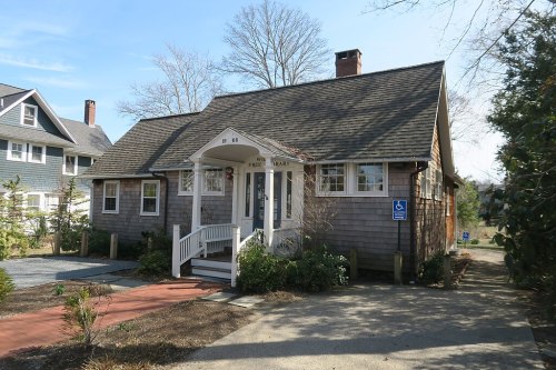 A charming, single-story wooden building with a sloped roof, labeled "Free Library," surrounded by greenery and a pathway.