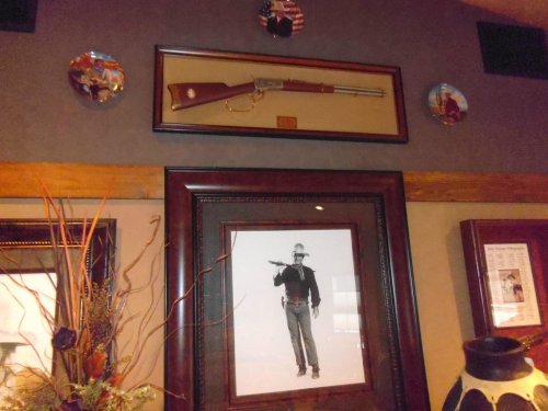 A framed vintage rifle hangs above a black-and-white photo of a cowboy, surrounded by decorative plates on the wall.