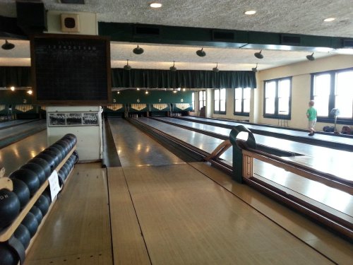 A bowling alley with wooden lanes, a scoreboard, and a few people in the background. Natural light from large windows.