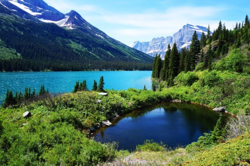 A serene landscape featuring a turquoise lake, lush greenery, and mountains under a clear blue sky.