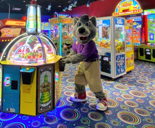 A costumed wolf character stands by a colorful arcade game in a vibrant game room filled with various machines.