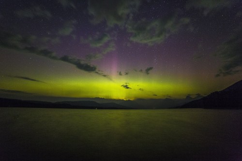 A serene night sky illuminated by green and purple auroras over a calm lake, with distant mountains silhouetted.