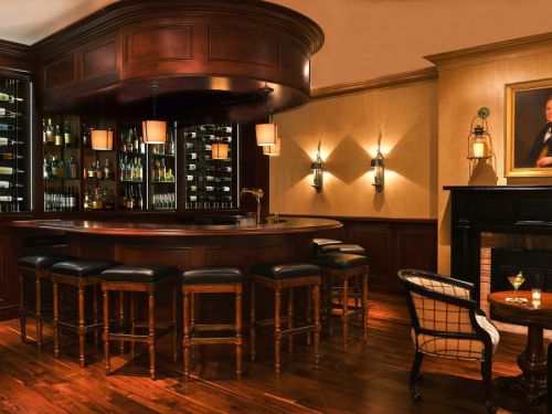 A cozy bar with a wooden counter, high stools, and a wine display, featuring warm lighting and elegant decor.