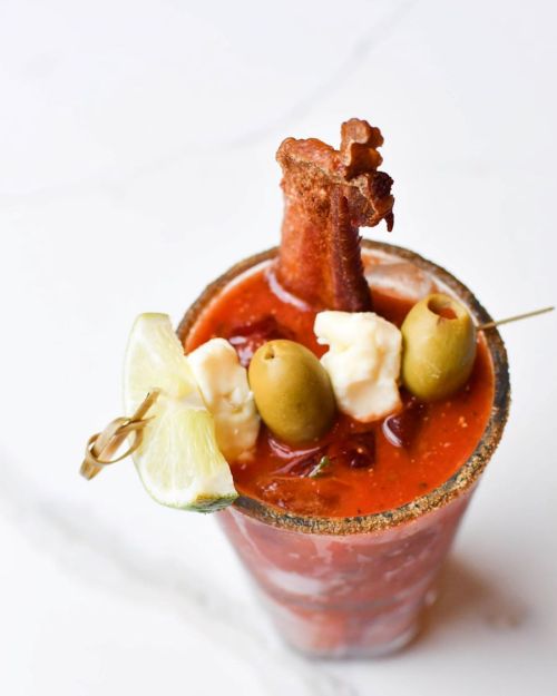 A vibrant Bloody Mary garnished with olives, cheese, lime, and crispy bacon in a salt-rimmed glass.