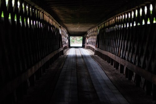 A dimly lit covered bridge with wooden beams, leading to a bright opening at the far end.