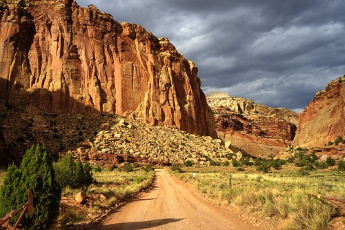 A dirt road winds through towering red rock formations under a cloudy sky, surrounded by sparse vegetation.