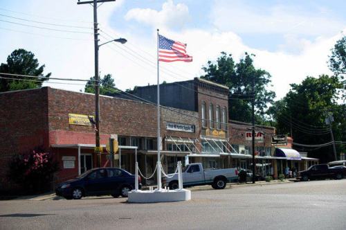 A small town street with brick buildings, an American flag, and parked cars under a clear blue sky.