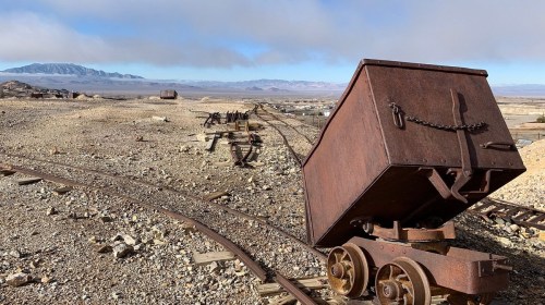 Rusty mining cart on tracks in a barren landscape with distant mountains under a cloudy sky.