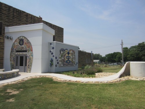 A colorful, mosaic-adorned building with a curved pathway and grassy area, set against a clear blue sky.