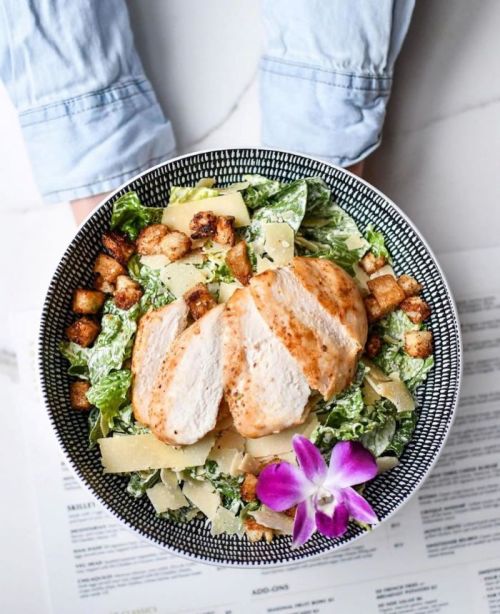 A bowl of Caesar salad topped with grilled chicken, croutons, Parmesan cheese, and a purple flower garnish.