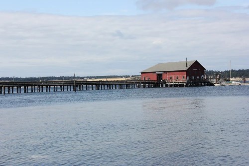 A red wooden building on a pier extending over calm water, with a cloudy sky in the background.
