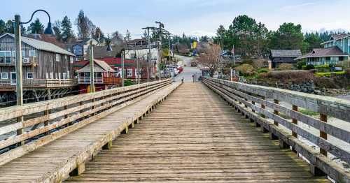 A wooden pier extends into a scenic waterfront village with buildings and trees lining the shore.