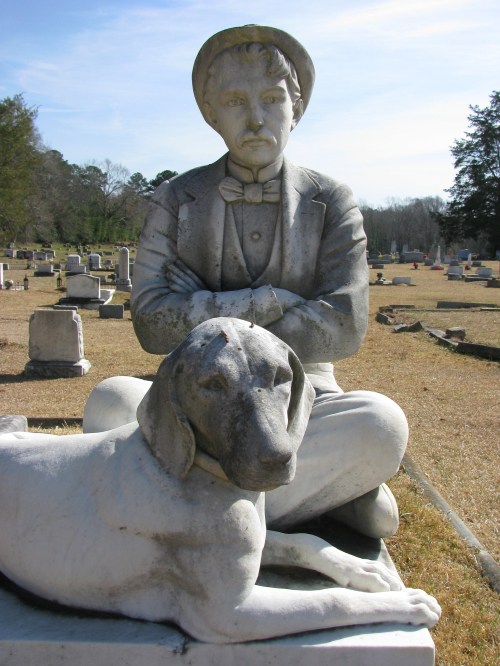 A marble statue of a seated man in a suit with a dog lying beside him, set in a cemetery with gravestones in the background.