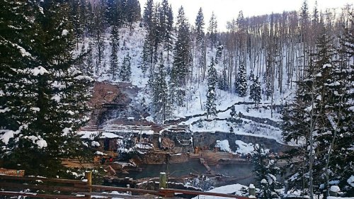 A snowy landscape featuring hot springs surrounded by evergreen trees and rocky terrain.
