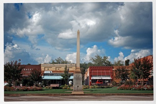 A town square featuring an obelisk surrounded by shops and blooming flowers under a cloudy sky.