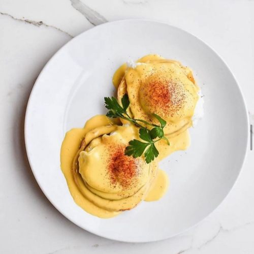 Two eggs Benedict on a white plate, topped with hollandaise sauce, paprika, and a sprig of parsley on a marble surface.