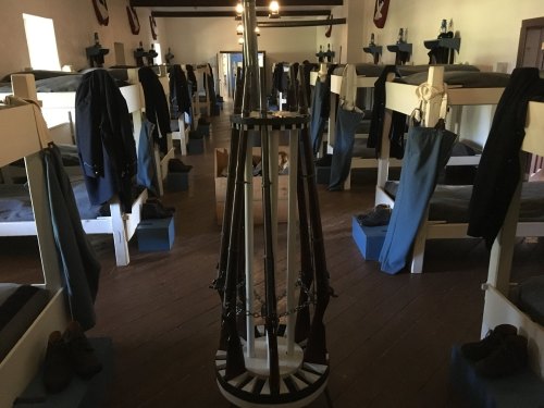 Interior of a barracks with bunk beds, clothing hanging, and a central stand holding rifles.