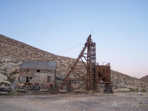 Abandoned mining structure with a tall wooden frame and a weathered building against a desert landscape at dusk.