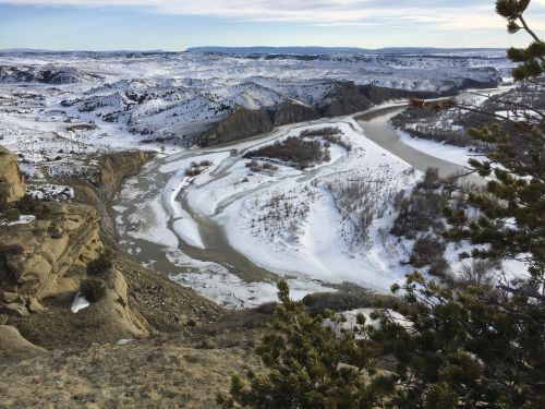 A winding river flows through a snowy landscape, surrounded by hills and sparse vegetation under a clear sky.