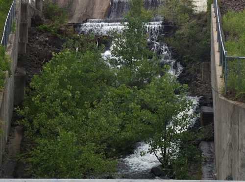 A small waterfall cascades over rocks, surrounded by greenery and concrete structures.