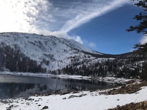A snowy mountain landscape with a calm lake reflecting the sky and surrounding trees under a partly cloudy sky.